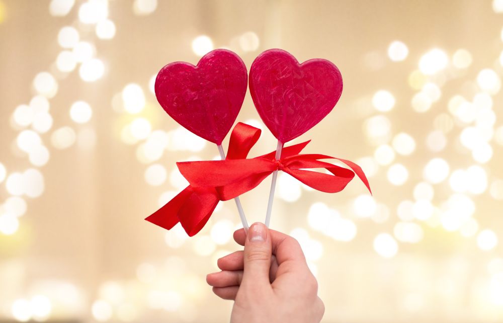 valentines day, sweets and romantic concept - close up of hand holding red heart shaped lollipops over festive lights background. close up of hand holding red heart shaped lollipop