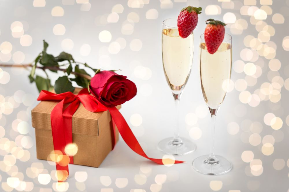 valentines day and holidays concept - two champagne glasses with strawberries and gift box with red rose over festive lights. two champagne glasses and gift with red rose