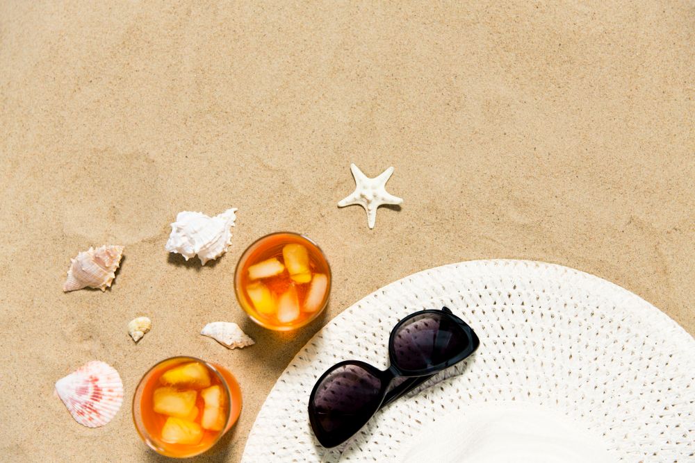vacation, travel and summer concept - two glasses of aperitif cocktails with ice cubes, sun hat, sunglasses and seashells on beach sand. cocktails, sun hat and sunglasses on beach sand