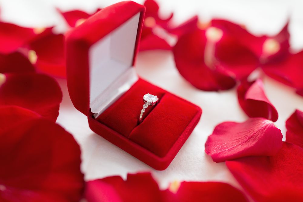 jewelry, proposal and romantic concept - diamond ring in red velvet gift box on bed sheet and rose petals. diamond ring in red velvet gift box on bed sheet