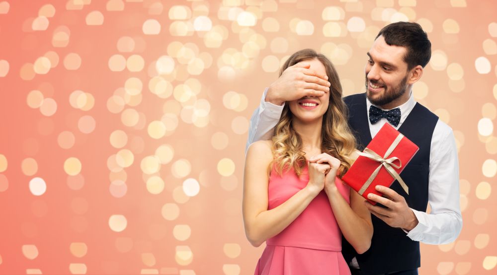valentines day, couple, relationships and people concept - happy man giving woman surprise present over living coral background with festive lights. happy man giving woman surprise present