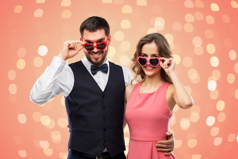 valentines day, love and people concept - happy couple in heart-shaped sunglasses over living coral background and festive lights. happy couple in heart-shaped sunglasses