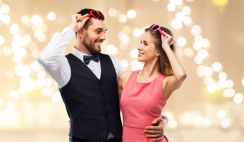 valentines day, love and people concept - happy couple in heart-shaped sunglasses over beige background with festive lights. happy couple in heart-shaped sunglasses