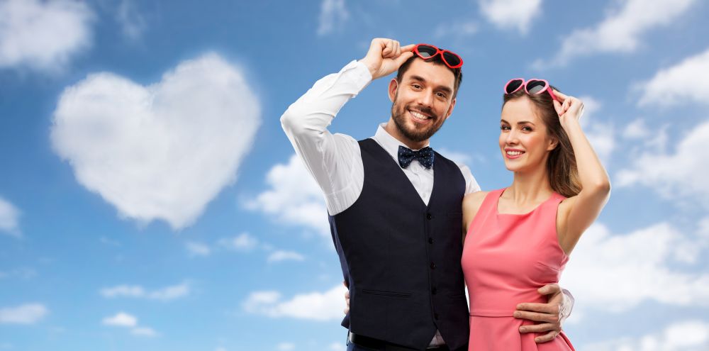 valentines day, love and people concept - happy couple in heart-shaped sunglasses over blue sky and heart shaped cloud background. happy couple in heart-shaped sunglasses