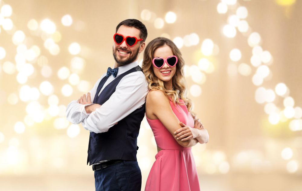 valentines day, love and people concept - happy couple in heart-shaped sunglasses over beige background with festive lights. happy couple in heart-shaped sunglasses
