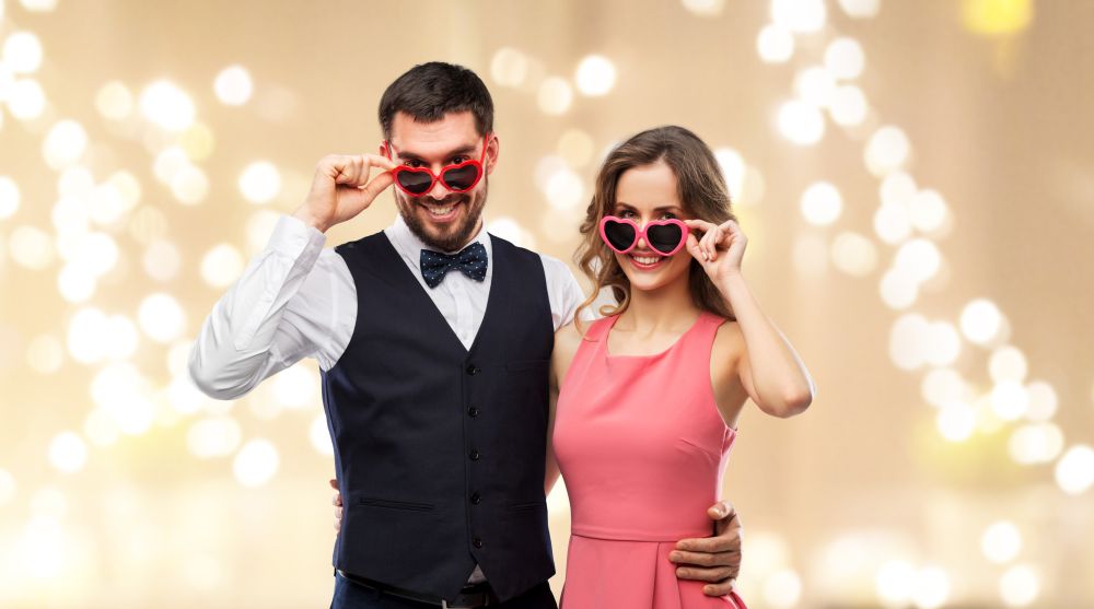 valentines day, love and people concept - happy couple in heart-shaped sunglasses over festive lights background. happy couple in heart-shaped sunglasses