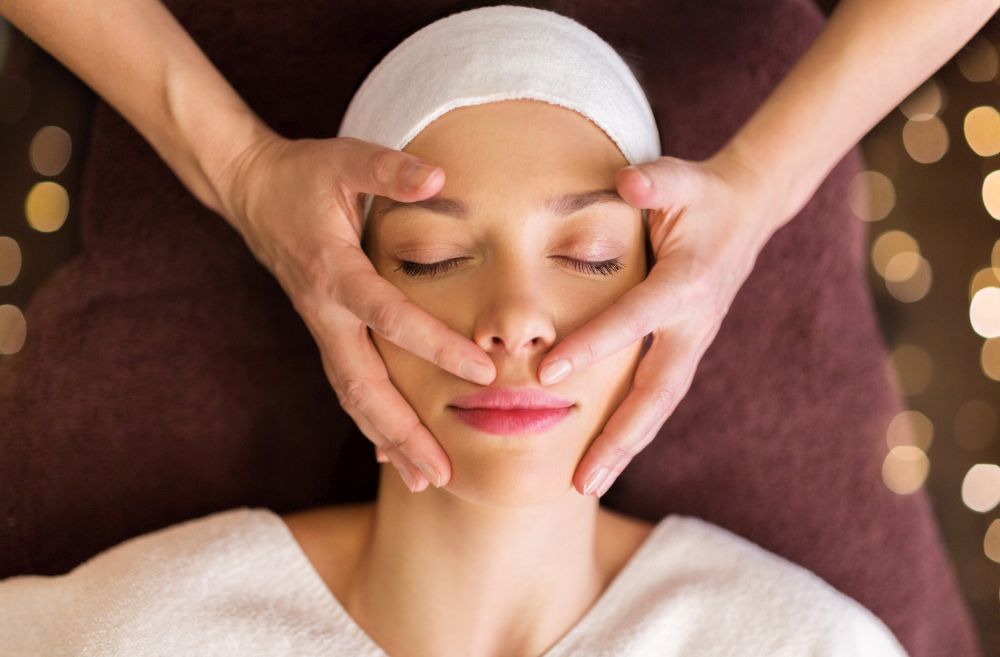people, beauty, lifestyle and relaxation concept - beautiful young woman lying with closed eyes and having face and head massage at spa. woman having face and head massage at spa