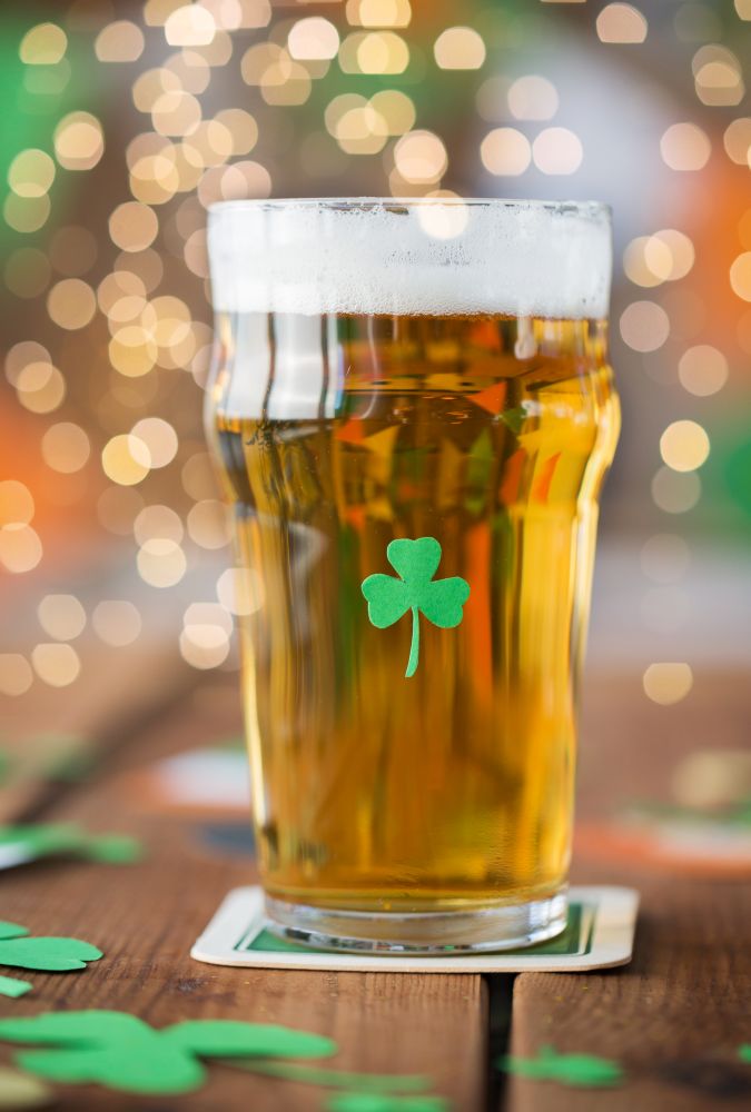 st patricks day, holidays and celebration concept - close up of glass of light beer with shamrock decoration on wooden table. close up of glass of beer with shamrock on table