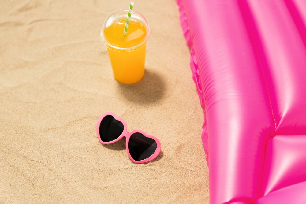 vacation and summer holidays concept - pink sunglasses, orange juice and swimming mattress on beach sand. sunglasses, juice and pool mattress on beach sand