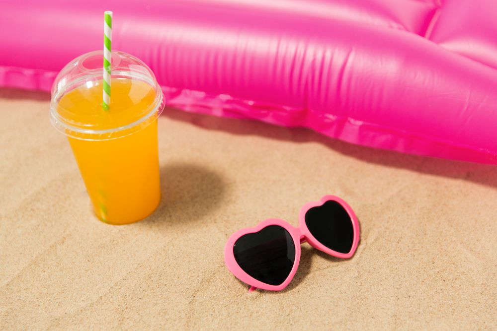vacation and summer holidays concept - pink sunglasses, orange juice and swimming mattress on beach sand. sunglasses, juice and pool mattress on beach sand