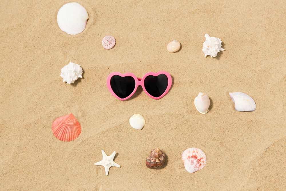 vacation, valentine&rsquo;s day and summer holidays concept - pink heart-shaped sunglasses and shells on beach sand. heart-shaped sunglasses and shells on beach sand