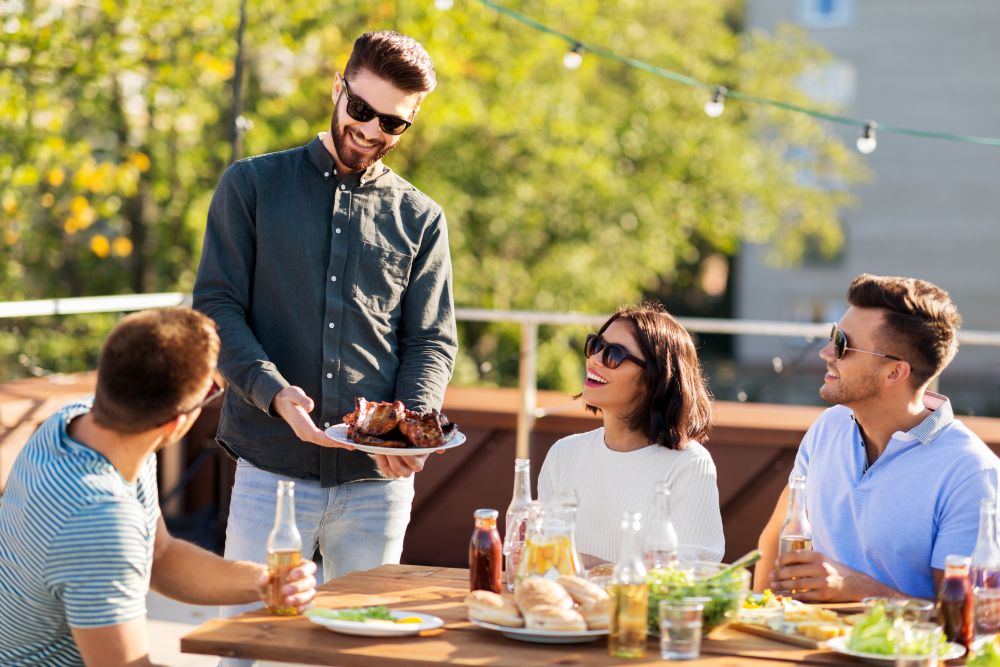leisure and people concept - happy party host offering meat to his friends at bbq on rooftop in summer. friends at bbq party on rooftop in summer