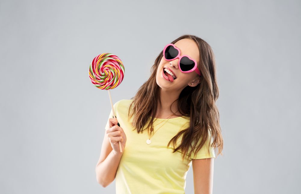 summer, valentine&rsquo;s day and people concept - smiling young woman or teenage girl in yellow t-shirt and heart-shaped sunglasses with lollipop over grey background. teenage girl in sunglasses with lollipop