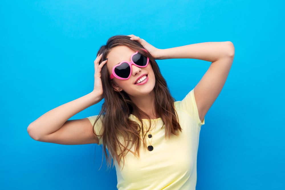 summer, valentine&rsquo;s day and people concept - smiling young woman or teenage girl in yellow t-shirt and heart-shaped sunglasses over bright blue background. teenage girl in heart-shaped sunglasses