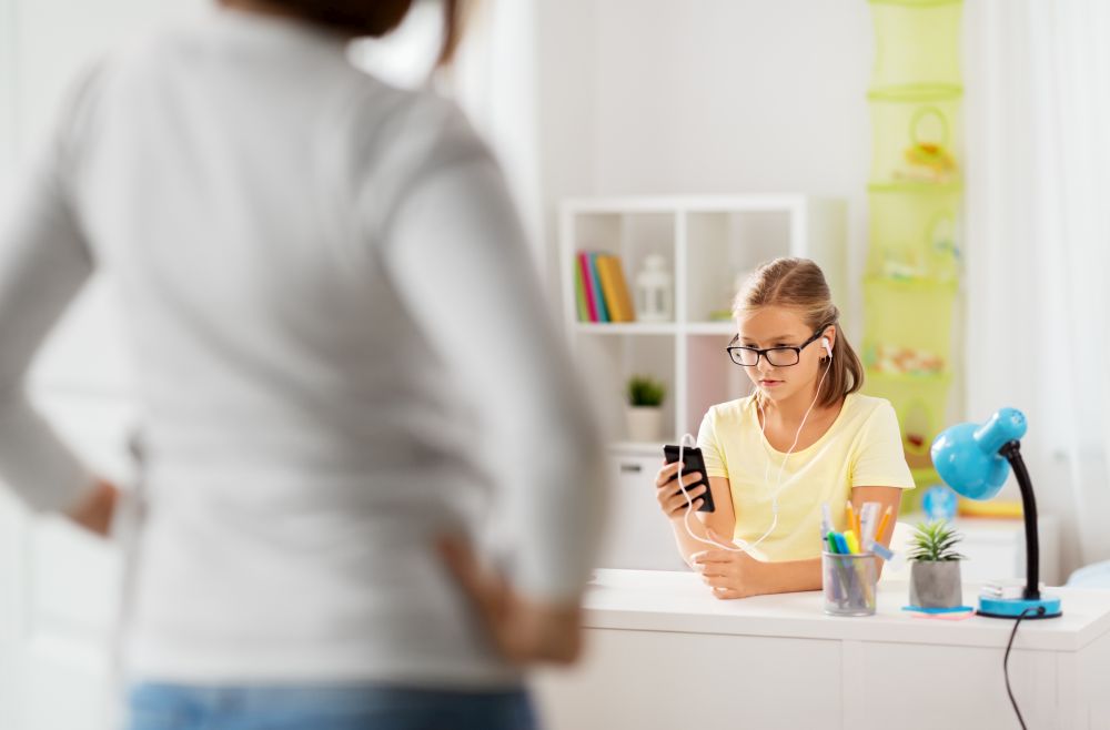 education, technology and school concept - daughter with smartphone distracting from homework and mother entering kids room. girl listening to music and mother entering room