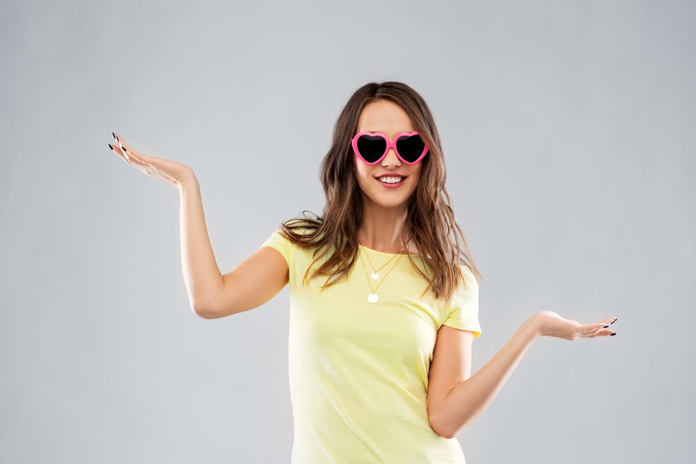 summer, valentine&rsquo;s day and people concept - smiling young woman or teenage girl in heart-shaped sunglasses holding something on palms over grey background. teenage girl in heart-shaped sunglasses
