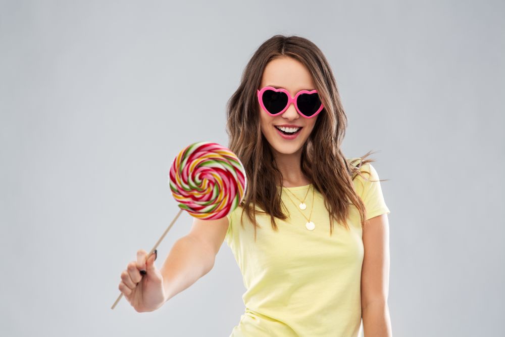 summer, valentine&rsquo;s day and people concept - smiling young woman or teenage girl in yellow t-shirt and heart-shaped sunglasses with lollipop over grey background. teenage girl in sunglasses with lollipop