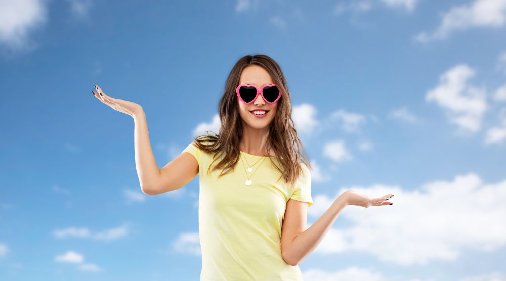 summer, valentine&rsquo;s day and people concept - smiling young woman or teenage girl in heart-shaped sunglasses holding something on palms over blue sky and clouds background. teenage girl in heart-shaped sunglasses