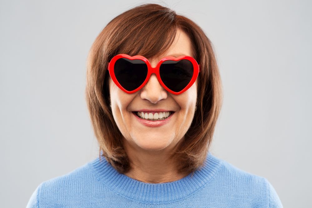 valentine&rsquo;s day, summer and old people concept - portrait of smiling senior woman in red heart-shaped sunglasses over grey background. smiling senior woman in heart-shaped sunglasses