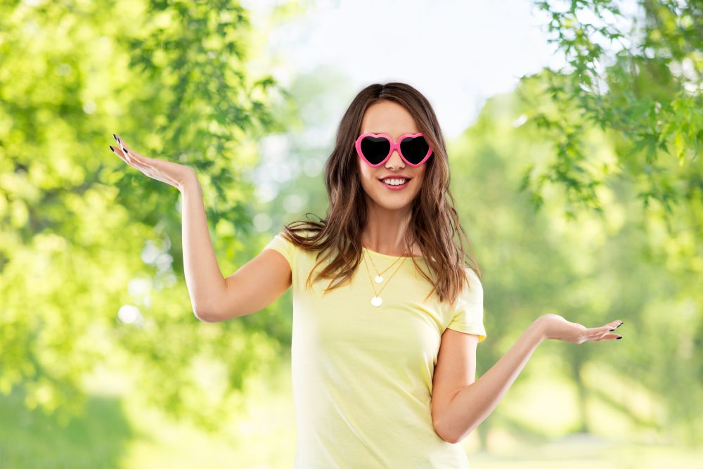 summer, valentine&rsquo;s day and people concept - smiling young woman or teenage girl in heart-shaped sunglasses holding something on palms over green natural background. teenage girl in heart-shaped sunglasses