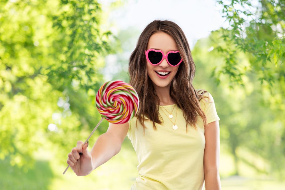 summer, valentine&rsquo;s day and people concept - smiling young woman or teenage girl in yellow t-shirt and heart-shaped sunglasses with lollipop over green natural background. teenage girl in sunglasses with lollipop