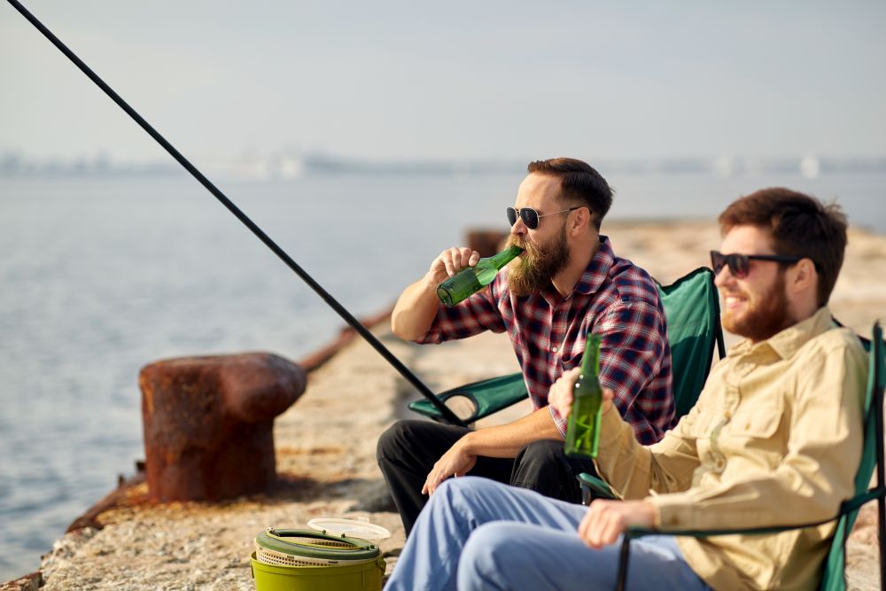 leisure and people concept - happy friends fishing and drinking beer on pier. happy friends fishing and drinking beer on pier