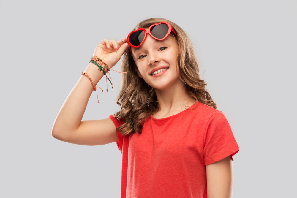 valentine&rsquo;s day, love and people concept - smiling pretty teenage girl wearing bangles on hand in red heart shaped sunglasses over grey background. happy teenage girl in red heart shaped sunglasses