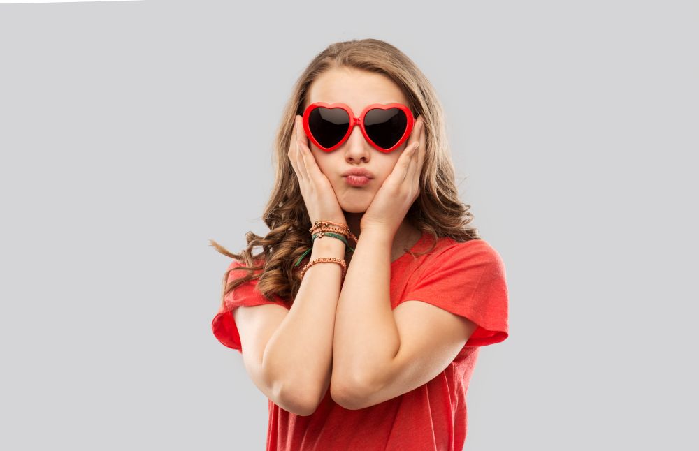 valentine&rsquo;s day, love and people concept - teenage girl in red heart shaped sunglasses touching her face and pouting over grey background. girl in red heart shaped sunglasses pouting