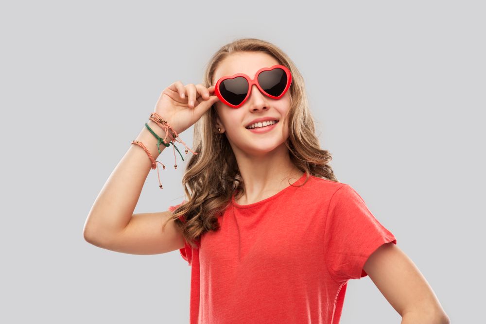 valentine&rsquo;s day, love and people concept - smiling pretty teenage girl wearing bangles on hand in red heart shaped sunglasses over grey background. happy teenage girl in red heart shaped sunglasses