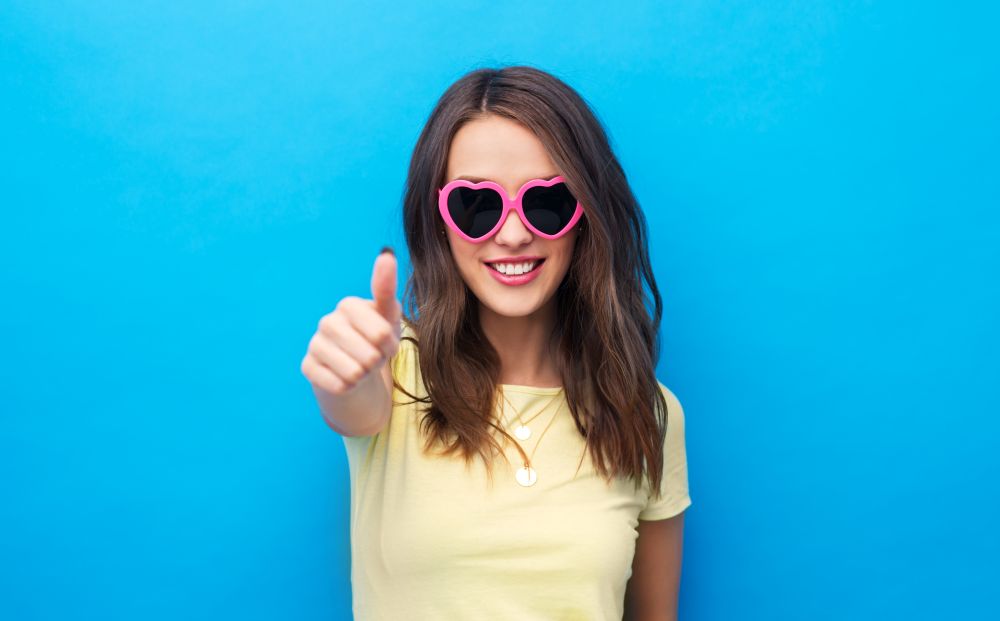 summer, valentine&rsquo;s day and people concept - smiling young woman or teenage girl in yellow t-shirt and heart-shaped sunglasses showing thumbs up over bright blue background. teenage girl in heart-shaped shades shows thumb up