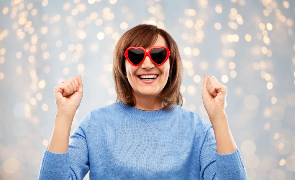 valentine&rsquo;s day, summer and old people concept - portrait of happy senior woman in red heart-shaped sunglasses celebrating triumph over festive lights background. smiling senior woman in heart-shaped sunglasses