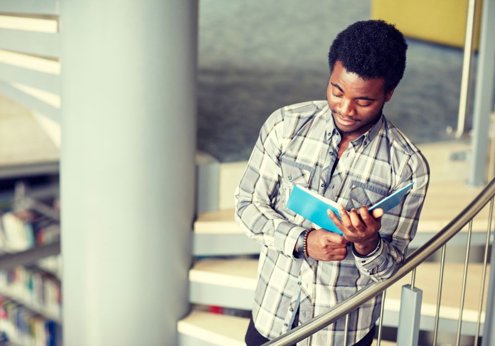 education, high school, university, learning and people concept - happy african american student boy or young man reading book on stairs at library. african student boy or man reading book at library