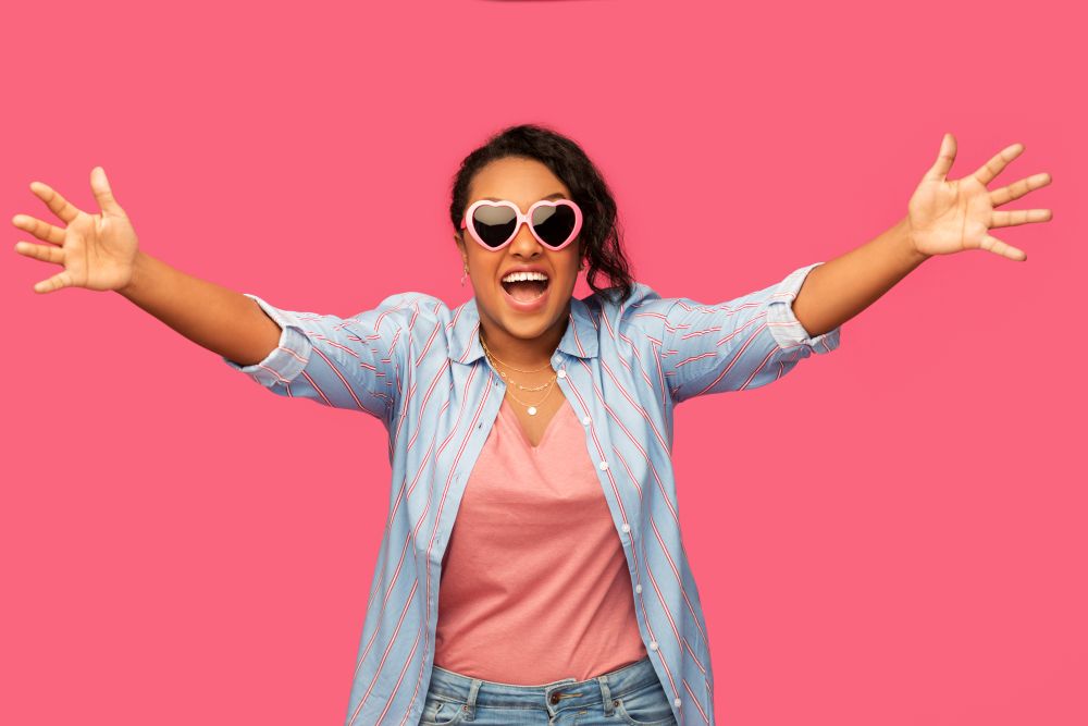 summer, valentine&rsquo;s day and emotion concept - happy african american young woman in heart-shaped sunglasses over pink background. african american woman in heart-shaped sunglasses