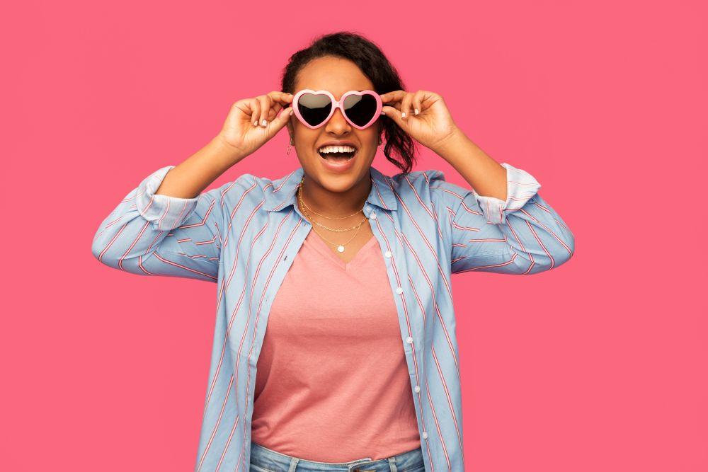 summer, valentine&rsquo;s day and eyewear concept - happy african american young woman in heart-shaped sunglasses over pink background. african american woman in heart-shaped sunglasses