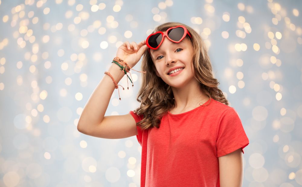 valentine&rsquo;s day, love and people concept - smiling pretty teenage girl wearing bangles on hand in red heart shaped sunglasses over festive lights background. happy teenage girl in red heart shaped sunglasses