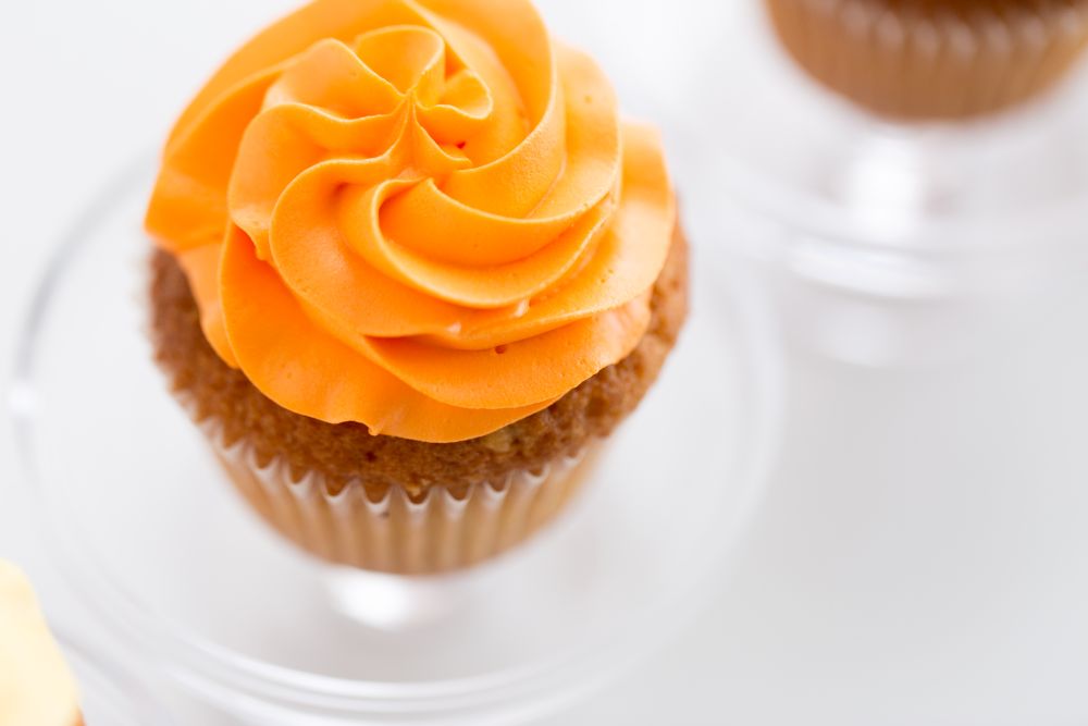 food, pastry and sweets concept - cupcake with orange buttercream frosting on glass confectionery stand over white background. cupcake with frosting on confectionery stand