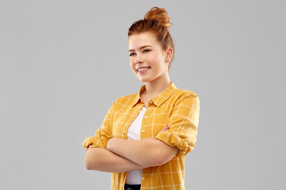 people concept - smiling red haired teenage girl in checkered shirt with crossed arms over grey background. smiling red haired teenage girl with crossed arms