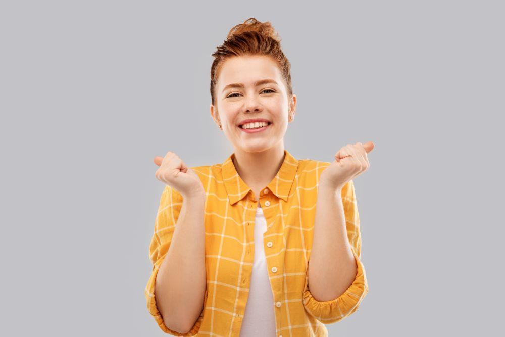 achievement, emotions and people concept - happy smiling red haired teenage girl in checkered shirt celebrating success over grey background. happy red haired teenage girl celebrating success