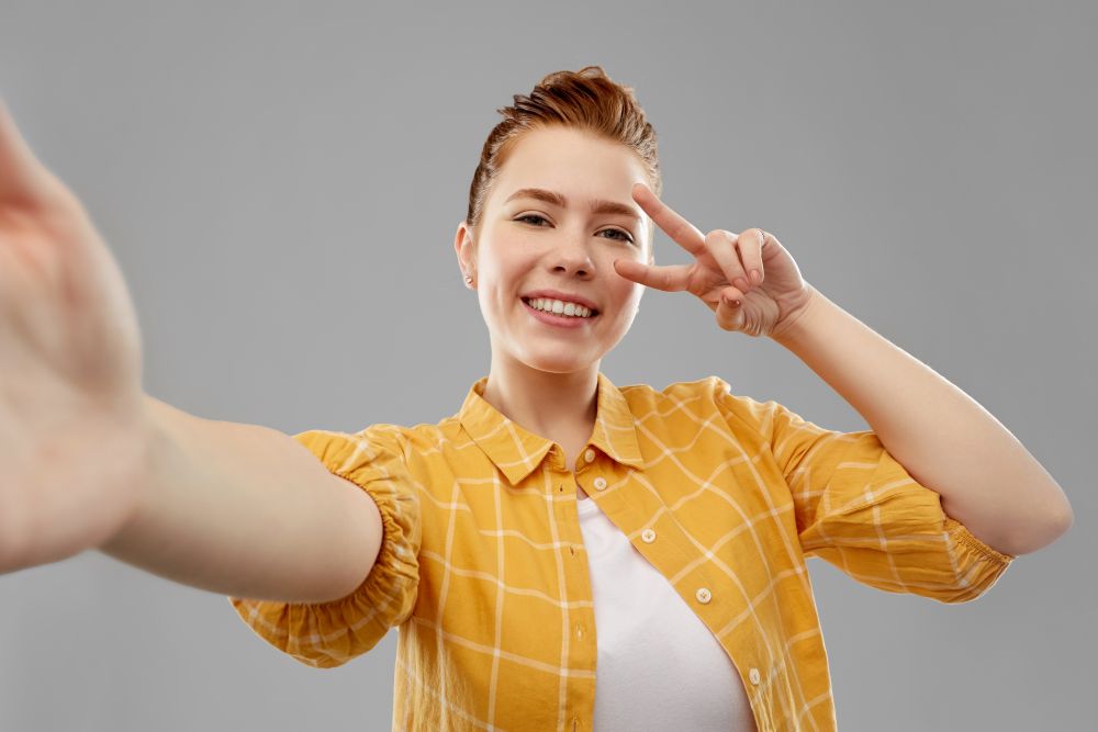 people concept - smiling red haired teenage girl in checkered shirt taking selfie and showing peace over grey background. redhead teenage girl taking selfie making peace