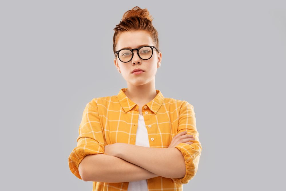 vision, education people concept - serious red haired teenage student girl in glasses and checkered shirt with crossed arms over grey background. redhead student girl in glasses with crossed arms