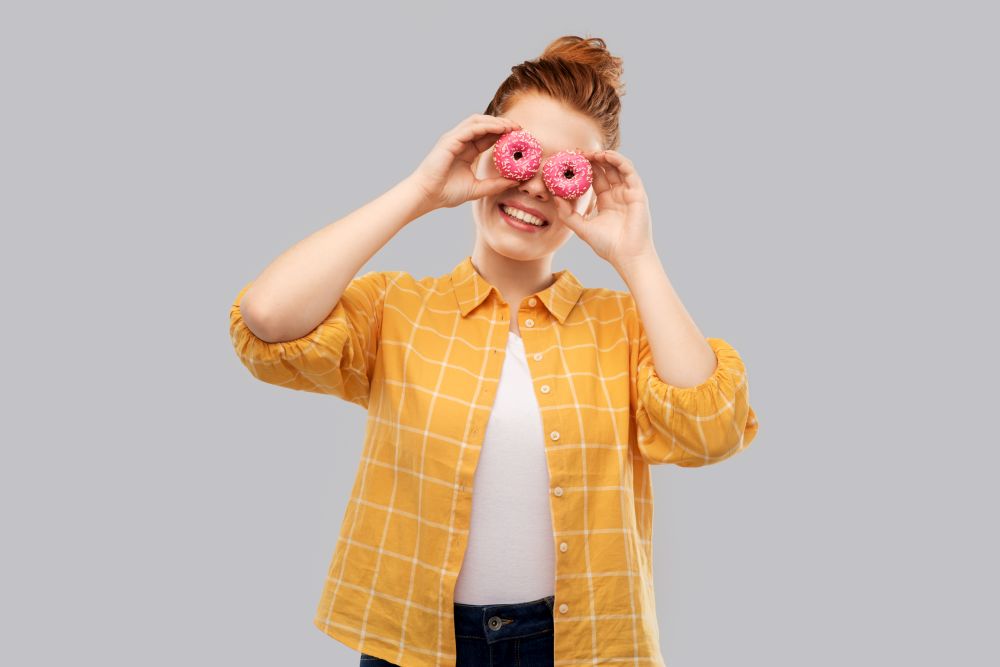 fast food and people concept - smiling red haired teenage girl in checkered shirt with donuts instead of eyes over grey background. funny teenage girl with donuts instead of eyes