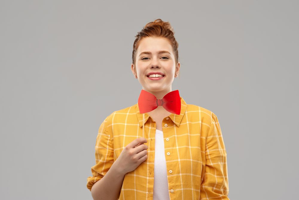 party props, photo booth and people concept - smiling red haired teenage girl in checkered shirt with big bowtie over grey background. smiling red haired teenage girl with big bowtie