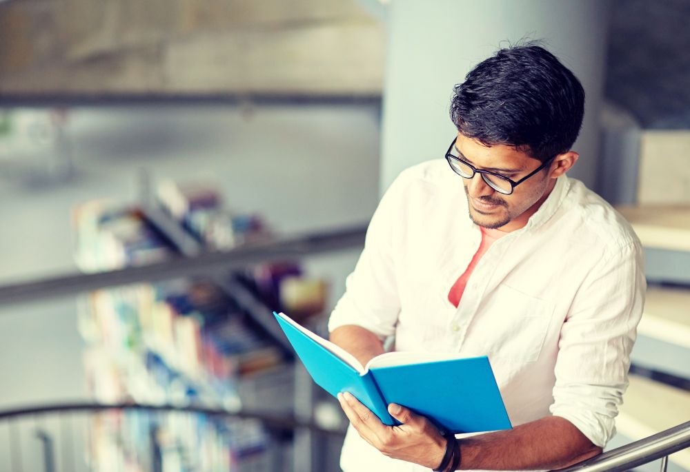 education, high school, university, learning and people concept - happy hindu student boy or young man reading book on stairs at library. hindu student boy or man reading book at library