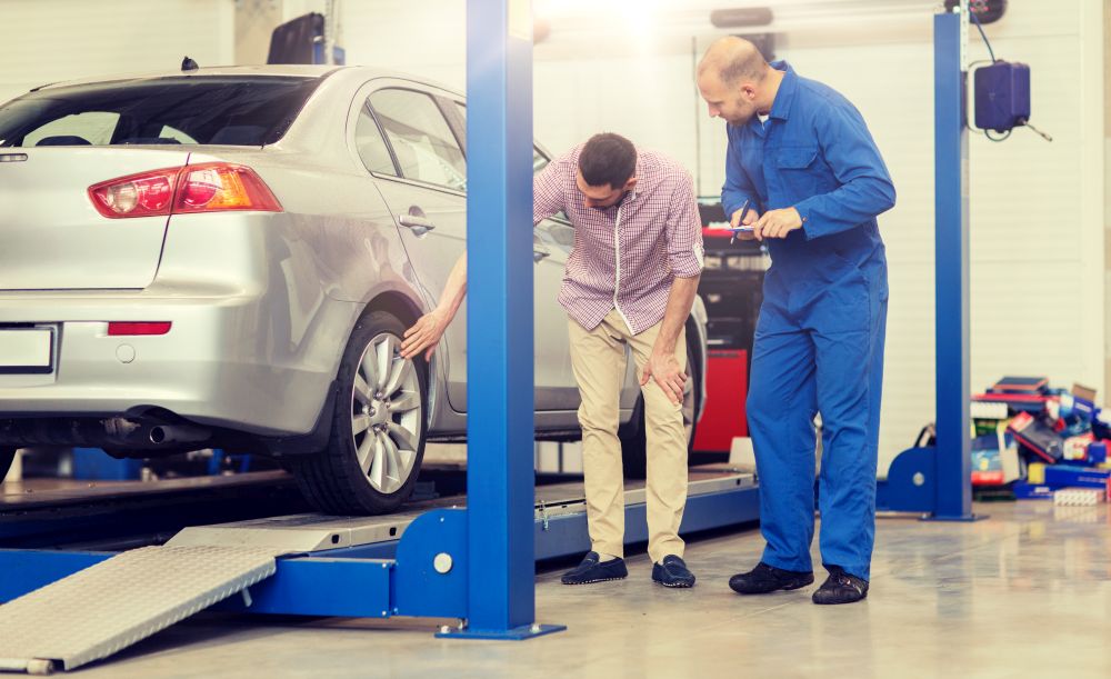 auto service, repair, maintenance and people concept - mechanic with clipboard and man or owner showing wheel at car shop. auto mechanic with clipboard and man at car shop