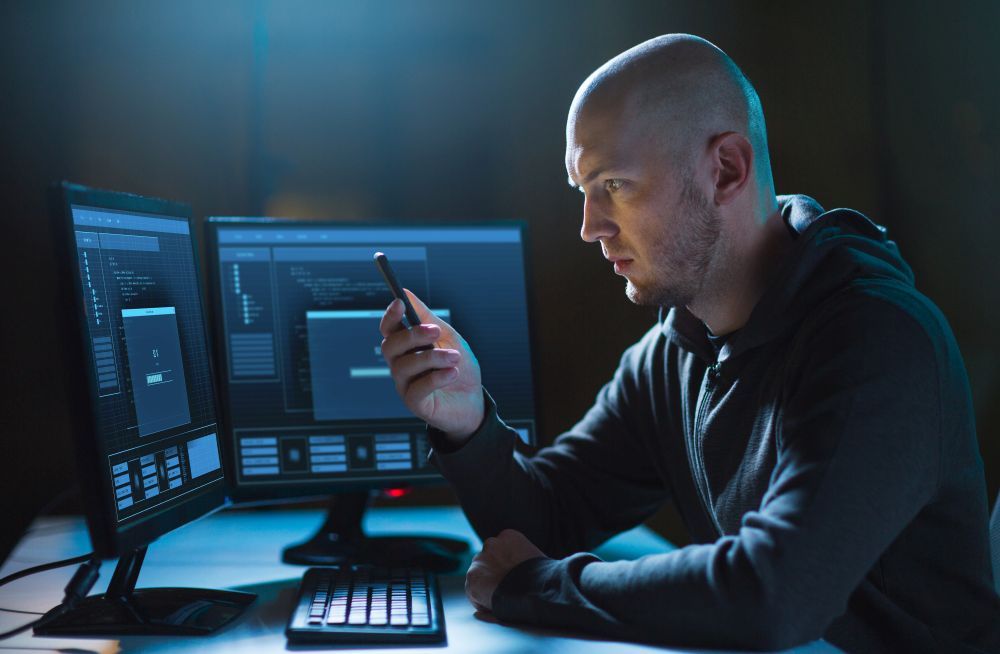 cybercrime, hacking and technology concept - male hacker with smartphone and progress loading bar on computer&rsquo;s screens in dark room. hacker with smartphone and computers in dark room