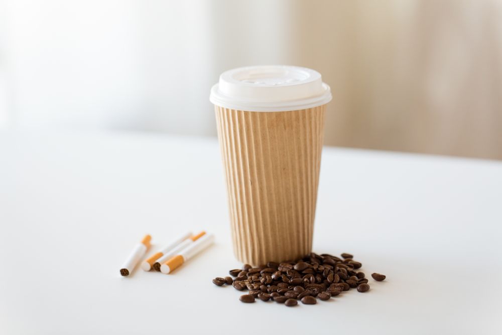 bad habits, addiction and unhealthy lifestyle concept - close up of cigarettes, takeaway coffee cup and roasted beans on white table. close up of cigarettes, coffee cup and beans