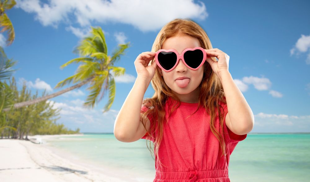 childhood, valentines day and summer concept - naughty red haired girl with heart shaped sunglasses showing tongue over tropical beach background in french polynesia. redhead girl in heart shaped sunglasses on beach