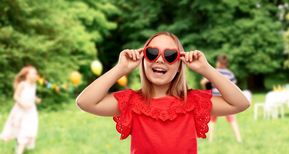 childhood, valentine&rsquo;s day and summer concept - happy preteen girl with heart shaped sunglasses over birthday party at park. happy girl with heart shaped sunglasses at party