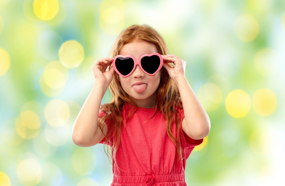 childhood, valentines day and summer concept - naughty red haired girl with heart shaped sunglasses showing tongue over green lights background. naughty red haired girl in heart shaped sunglasses