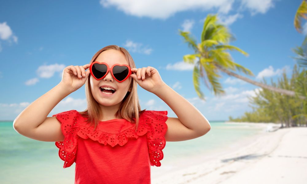 childhood, valentine&rsquo;s day and summer concept - happy preteen girl with heart shaped sunglasses over tropical beach background in french polynesia. happy girl with heart shaped sunglasses on beach